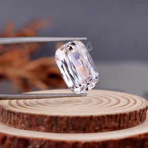 May include: A clear, colorless, cushion-cut gemstone with a faceted surface that reflects light. The stone is held by metal tweezers, displaying a rectangular shape with rounded corners and a high degree of transparency and brilliance.