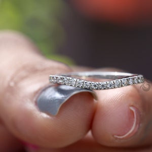May include: A silver-toned curved band ring featuring a row of small, clear, round-cut stones set along the top edge. The metal has a polished finish, and the band follows a gentle arc shape, showcasing a series of sparkling stones embedded in the metal.