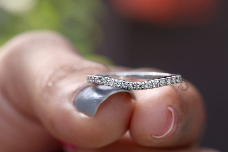 May include: A silver-toned curved band ring featuring a row of small, clear, round-cut stones set along the top edge. The metal has a polished finish, and the band follows a gentle arc shape, showcasing a series of sparkling stones embedded in the metal.