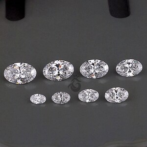 May include: A collection of eight clear, oval-cut gemstones arranged in two rows. The top row features four larger stones, while the bottom row contains four smaller stones. Each gemstone exhibits multiple facets that reflect light, creating a brilliant, sparkling appearance against a dark gray background.