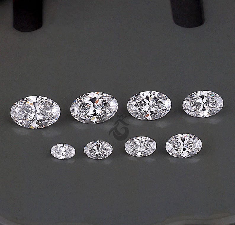 May include: A collection of eight clear, oval-cut gemstones arranged in two rows. The top row features four larger stones, while the bottom row contains four smaller stones. Each gemstone exhibits multiple facets that reflect light, creating a brilliant, sparkling appearance against a dark gray background.