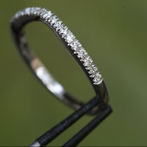 May include: A thin, polished silver-toned metal ring band featuring a row of small, clear, round-cut stones set along the top half of the band. The ring is held by a black tool, showcasing the reflective surface and the sparkling stones.