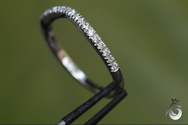 May include: A thin, polished silver-toned metal ring band featuring a row of small, clear, round-cut stones set along the top half of the band. The ring is held by a black tool, showcasing the reflective surface and the sparkling stones.