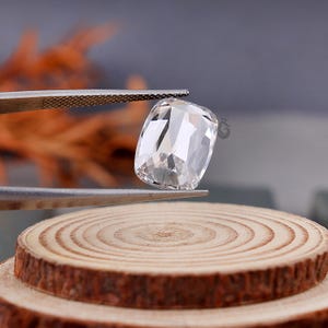 May include: A clear, cushion-cut gemstone held by metal tweezers. The stone features multiple facets that reflect light, creating a brilliant, transparent appearance. The gemstone is positioned above a circular, light-colored wooden slice.