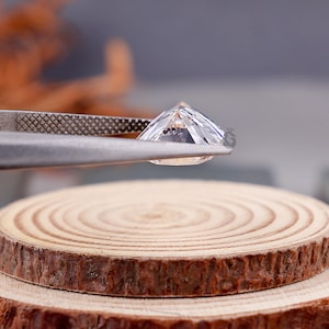 May include: A clear, faceted gemstone held by metal tweezers. The stone features a pointed crown and a flat table, displaying internal light reflections and sharp edges. The gemstone is positioned above a circular, light-colored wooden slice.