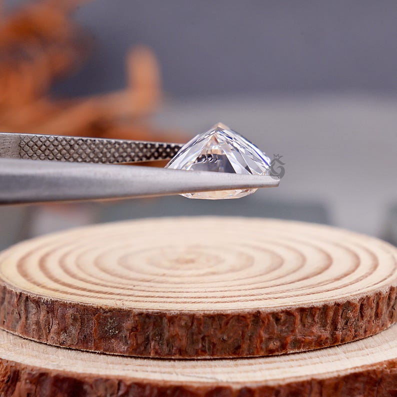 May include: A clear, faceted gemstone held by metal tweezers. The stone features a pointed crown and a flat table, displaying internal light reflections and sharp edges. The gemstone is positioned above a circular, light-colored wooden slice.