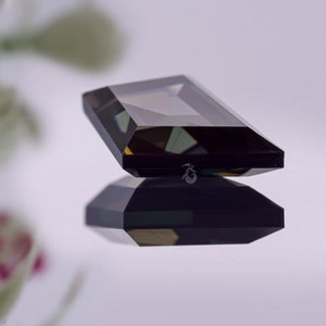 May include: A rectangular, emerald-cut gemstone with a deep, dark brown hue and faceted surfaces that reflect light. The stone is positioned above its own clear reflection, and a small, stylized crown-like logo is visible on the side of the gem.