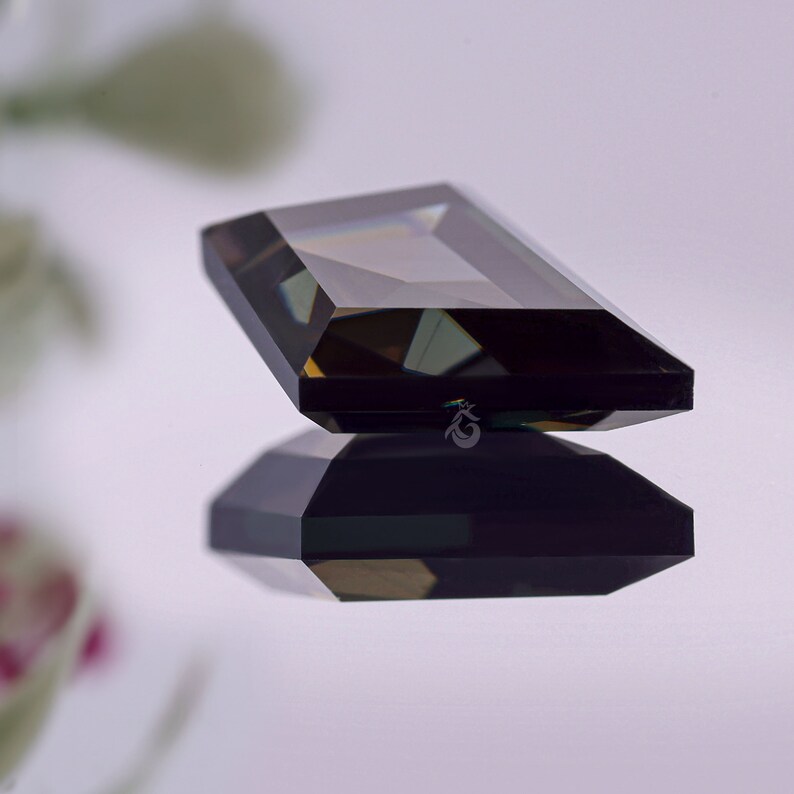 May include: A rectangular, emerald-cut gemstone with a deep, dark brown hue and faceted surfaces that reflect light. The stone is positioned above its own clear reflection, and a small, stylized crown-like logo is visible on the side of the gem.