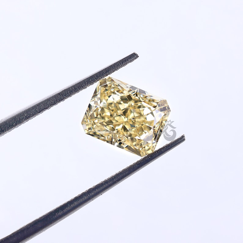 May include: A radiant-cut yellow gemstone held by dark metal tweezers. The stone features a rectangular shape with numerous triangular and trapezoidal facets that create a complex, sparkling pattern of light and shadow across its surface.