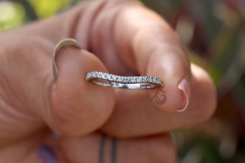 May include: A silver-toned curved band ring featuring a row of small, clear, round-cut stones set along the top surface. The metal band has a polished finish and follows a gentle wave-like contour, showcasing a series of sparkling accents.