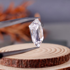 May include: A clear, faceted gemstone with a hexagonal elongated shape held by dark metal tweezers. The stone features multiple triangular and trapezoidal facets that reflect light, creating a bright, transparent appearance with sharp edges.