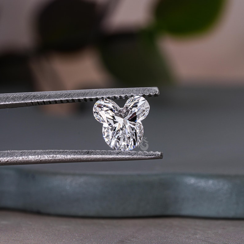 May include: A clear, heart-shaped diamond held in tweezers. The diamond has a unique, Mickey Mouse-like shape.