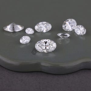 May include: A collection of eight clear, faceted, oval-shaped gemstones arranged in a cluster. Each stone features multiple sharp, geometric facets that reflect light, creating a brilliant, sparkling appearance across the various sizes shown.