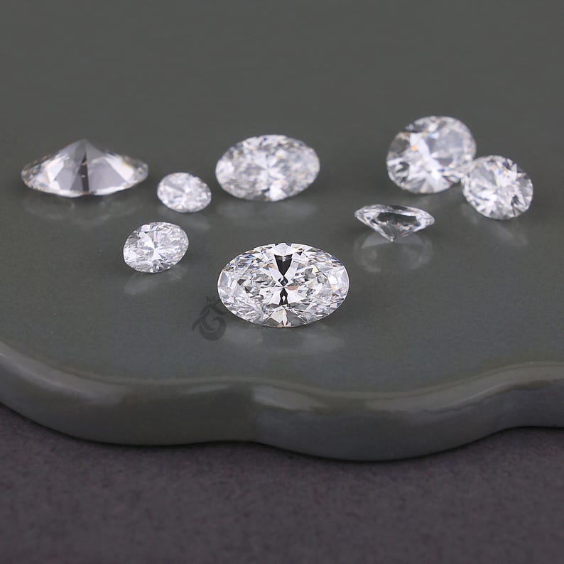 May include: A collection of eight clear, faceted, oval-shaped gemstones arranged in a cluster. Each stone features multiple sharp, geometric facets that reflect light, creating a brilliant, sparkling appearance across the various sizes shown.
