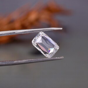 May include: A clear, colourless, rectangular gemstone with a radiant cut, held by a pair of metal tweezers. The stone features multiple facets that reflect light, creating sharp geometric patterns and internal highlights across its surface.