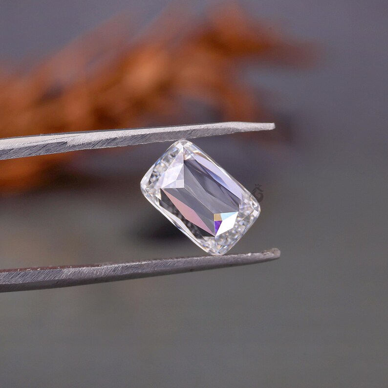 May include: A clear, colourless, rectangular gemstone with a radiant cut, held by a pair of metal tweezers. The stone features multiple facets that reflect light, creating sharp geometric patterns and internal highlights across its surface.
