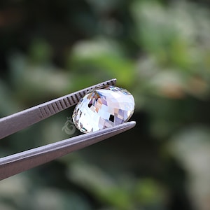 May include: A clear, round, faceted gemstone held by metal tweezers. The stone features a brilliant cut with multiple triangular facets that reflect light, creating a sparkling appearance. A small logo is visible on the side of the stone.