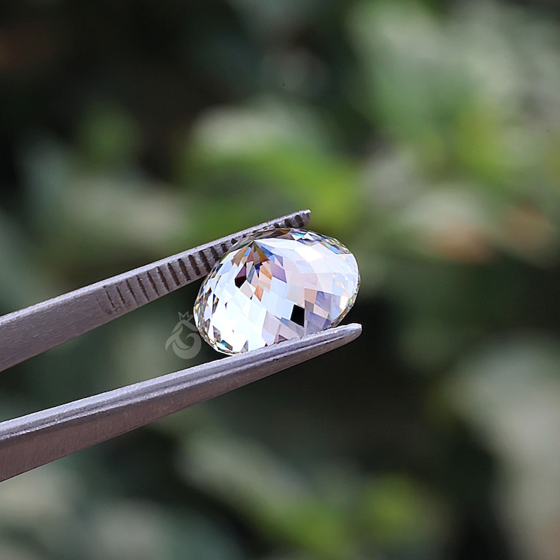 May include: A clear, round, faceted gemstone held by metal tweezers. The stone features a brilliant cut with multiple triangular facets that reflect light, creating a sparkling appearance. A small logo is visible on the side of the stone.