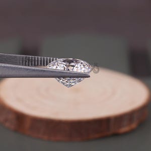 May include: A clear, round, brilliant-cut gemstone held by metal tweezers. The stone features multiple facets that reflect light, creating a sparkling appearance. The gemstone is transparent and colorless, showcasing a precise geometric cut.