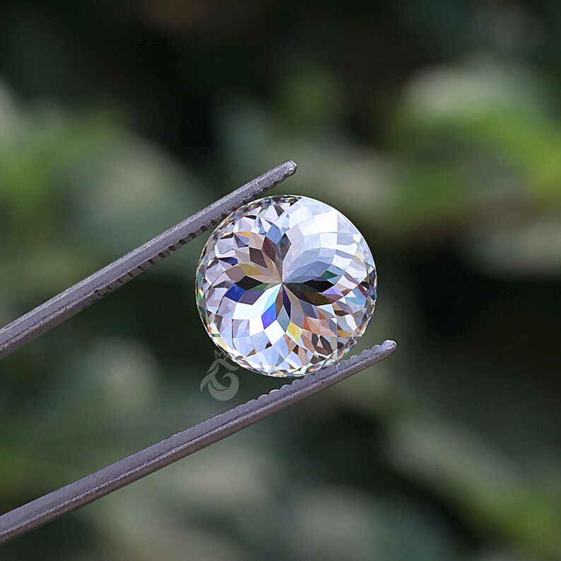 May include: A round, clear, faceted gemstone held by metal tweezers. The stone features a complex, symmetrical cut that creates a star-like pattern of light reflections and internal refractions, displaying subtle rainbow-colored flashes.