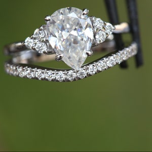 May include: A silver-toned engagement ring set featuring a pear-cut center stone flanked by three small round stones on each side, paired with a curved wedding band encrusted with a row of small round stones along the entire top edge.