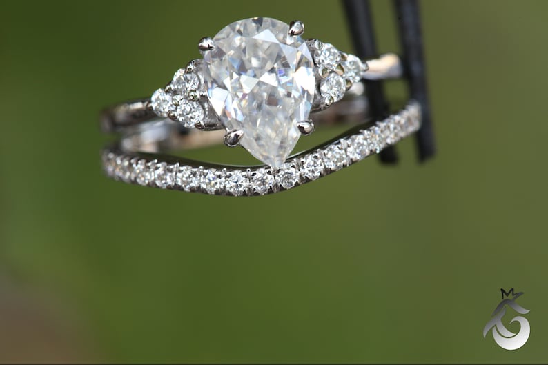 May include: A silver-toned engagement ring set featuring a pear-cut center stone flanked by three small round stones on each side, paired with a curved wedding band encrusted with a row of small round stones along the entire top edge.