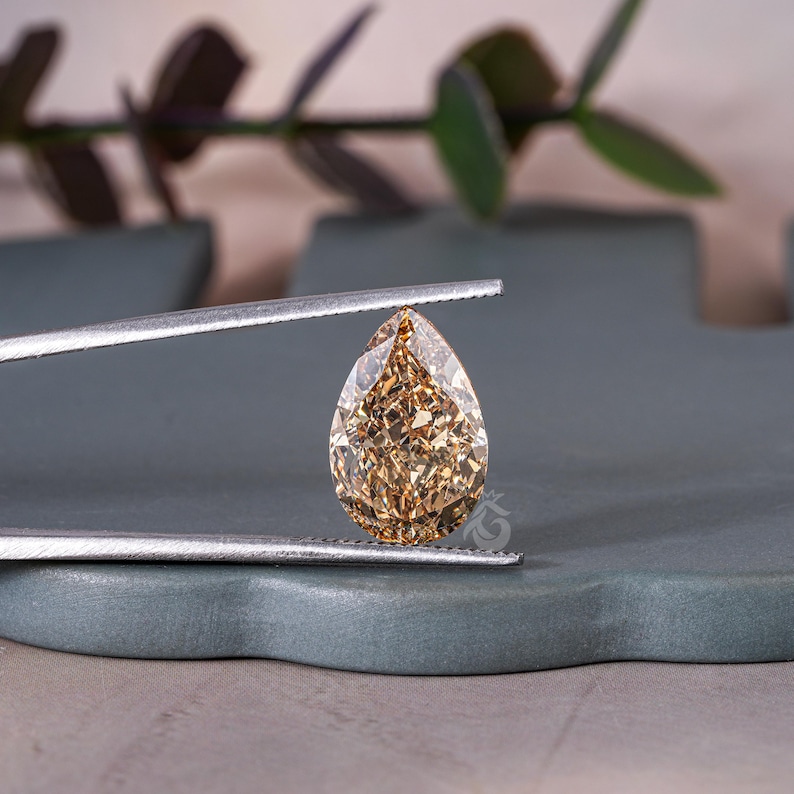 May include: A pear-shaped champagne-colored gemstone with a faceted surface held by silver-toned metal tweezers. The stone exhibits a warm, brownish-gold hue with intricate light-reflecting cuts that create a brilliant, sparkling appearance.