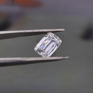 May include: A clear, colourless, emerald-cut gemstone held by a pair of metal tweezers. The stone features a rectangular shape with faceted edges that reflect light, creating a brilliant, transparent appearance with sharp, clean lines throughout.