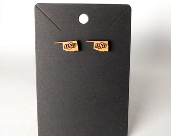 May include: A pair of wooden earrings shaped like the state of Oklahoma with the letters "OSU" in the center. The earrings are on a black display card.