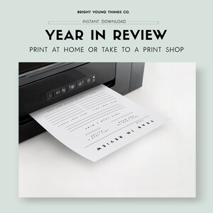 May include: A black and white printable year in review planner sheet is being printed from a black printer. The sheet has lines for writing and the text "YEAR IN REVIEW" is printed at the top and bottom of the sheet.
