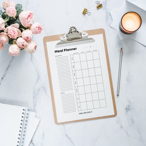 May include: A white meal planner on a clipboard with a brown backing. The planner has sections for shopping, notes, and a weekly meal plan with columns for breakfast, lunch, dinner, and snacks.
