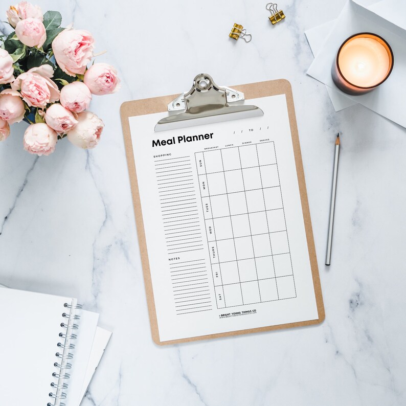 May include: A white meal planner on a clipboard with a brown backing. The planner has sections for shopping, notes, and a weekly meal plan with columns for breakfast, lunch, dinner, and snacks.