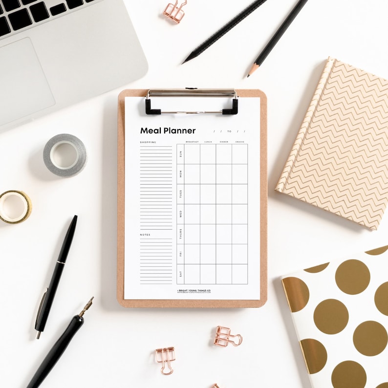 May include: A white meal planner on a clipboard with a brown wooden frame. The planner has sections for shopping, notes, breakfast, lunch, dinner, and snacks. The planner is on a white surface with a laptop, pencils, and decorative notebooks.