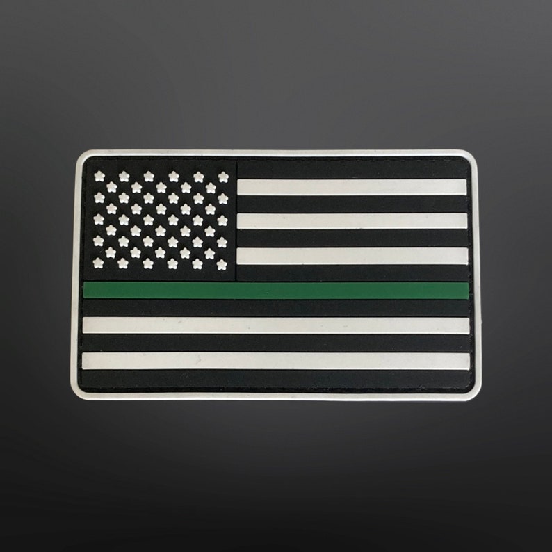 Op de afbeelding: Een zwart-wit Amerikaanse vlag patch met een groene streep in het midden. De vlag is gemaakt van een getextureerd materiaal en heeft een witte rand.