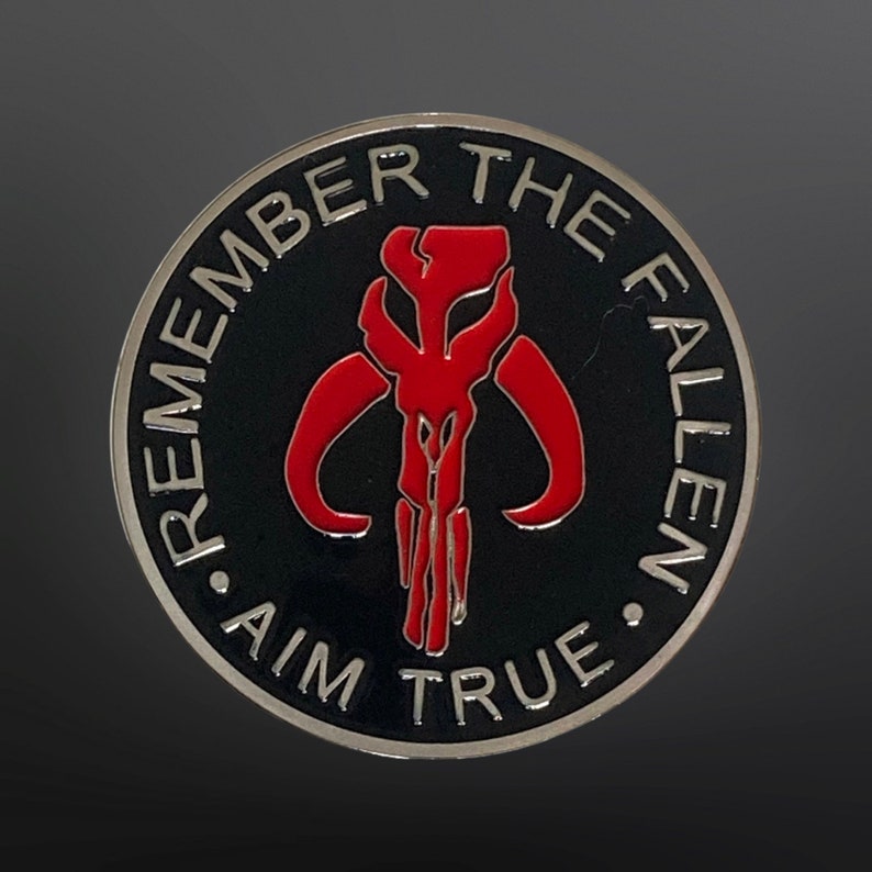 May include: A black and silver round pin with the Mandalorian symbol in red. The text around the symbol reads "Remember the Fallen. Aim True."
