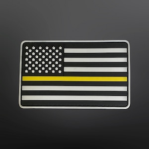 Puede incluir: Un parche de bandera estadounidense en blanco y negro con una franja amarilla en el medio. El parche está hecho de un material texturizado y tiene un borde blanco.