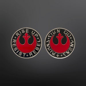May include: Two black and silver circular pins with a red rebel alliance symbol. The text "Rise Up Rebel" is written around the symbol on one pin. The other pin has the symbol surrounded by a circle of text in a foreign language.