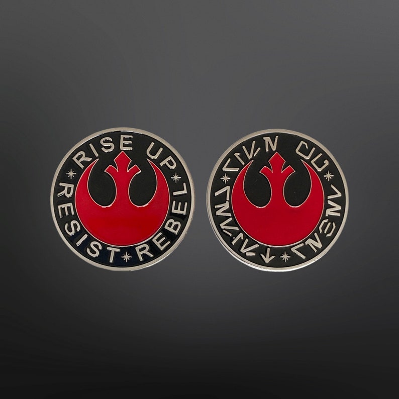 May include: Two black and silver circular pins with a red rebel alliance symbol. The text "Rise Up Rebel" is written around the symbol on one pin. The other pin has the symbol surrounded by a circle of text in a foreign language.