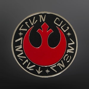 May include: A black and silver pin with a red Rebel Alliance symbol and the text "REBEL ALLIANCE" in Aurebesh script around the edge.