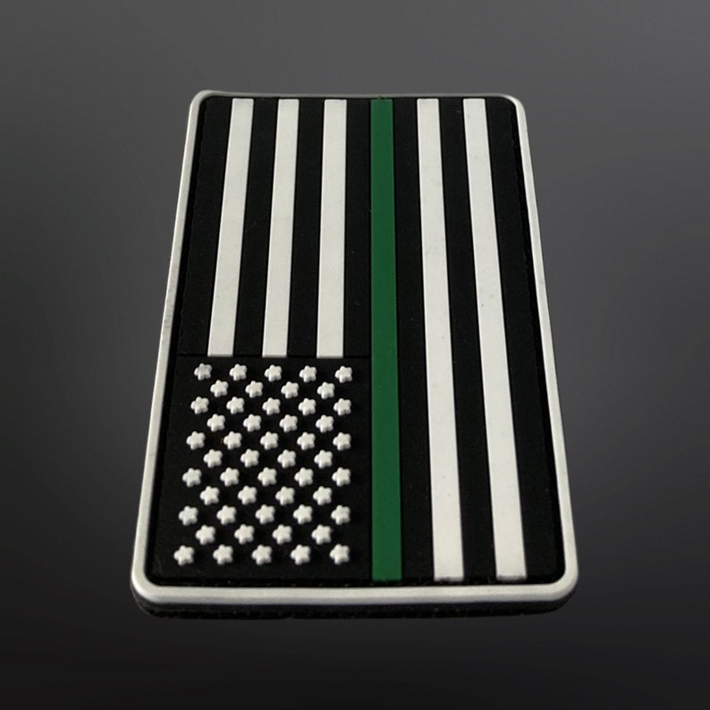 Op de afbeelding: Een zwart-witte Amerikaanse vlagpatch met een groene streep in het midden. De patch is gemaakt van duurzaam materiaal en is perfect om een ​​patriotische touch aan je bezittingen toe te voegen.