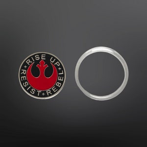May include: A silver and black round pin with the Rebel Alliance symbol and the text "*RESIST* RISE UP* REBEL*" in a circle around the symbol. The symbol is red.