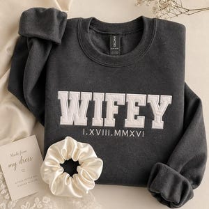 May include: Dark gray sweatshirt with the word "WIFEY" in white, block letters. Below the word is the roman numeral "I.XVIII.MMXVI". A white satin scrunchie and a card that reads "Made from my dress" are also in the image.