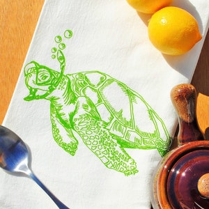 Puede incluir: Una toalla de cocina blanca con un dibujo de l&iacute;nea verde de una tortuga marina que lleva una m&aacute;scara de buceo y un snorkel. La tortuga est&aacute; rodeada de burbujas. La toalla est&aacute; sobre una superficie de madera con limones y una tetera de cer&aacute;mica marr&oacute;n.