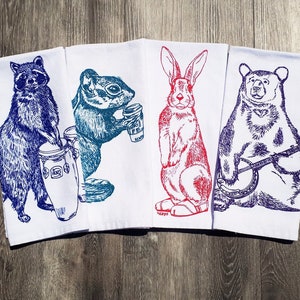 Peut inclure: Ensemble de quatre torchons de cuisine avec des illustrations colorées d'un raton laveur jouant du tambour, d'un écureuil tenant une tasse de café, d'un lapin et d'un ours jouant du banjo. Les torchons sont blancs avec de l'encre bleue, rouge et violette.