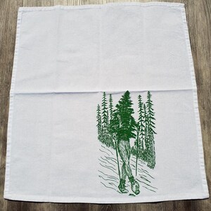 Pode incluir: Toalha de cozinha branca com uma ilustra&ccedil;&atilde;o verde de uma pessoa a fazer caminhadas por uma floresta. A pessoa est&aacute; a usar botas de caminhada e a levar bast&otilde;es de caminhada. As &aacute;rvores s&atilde;o altas e esguias.
