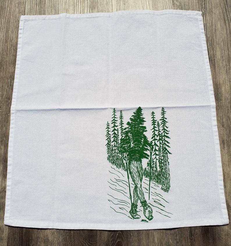 Pode incluir: Toalha de cozinha branca com uma ilustra&ccedil;&atilde;o verde de uma pessoa a fazer caminhadas por uma floresta. A pessoa est&aacute; a usar botas de caminhada e a levar bast&otilde;es de caminhada. As &aacute;rvores s&atilde;o altas e esguias.