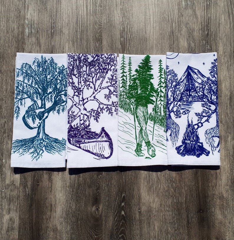 Pode incluir: Conjunto de quatro toalhas de cozinha com ilustra&ccedil;&otilde;es azuis e brancas de cenas da natureza. Os desenhos incluem uma &aacute;rvore com um viol&atilde;o, uma canoa, uma pessoa na floresta e uma fogueira.