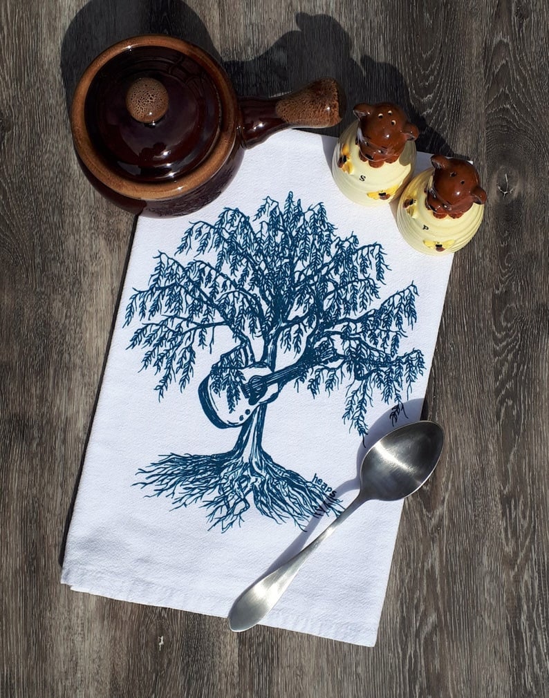 May include: White kitchen towel with a blue illustration of a tree with a guitar in its branches. The tree has roots and the text "Heavens" is visible at the bottom of the image.
