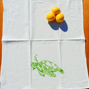 Puede incluir: Toalla de cocina blanca con una ilustraci&oacute;n verde de una tortuga marina con burbujas que salen de su nariz. Tres limones amarillos est&aacute;n en la parte superior de la toalla.