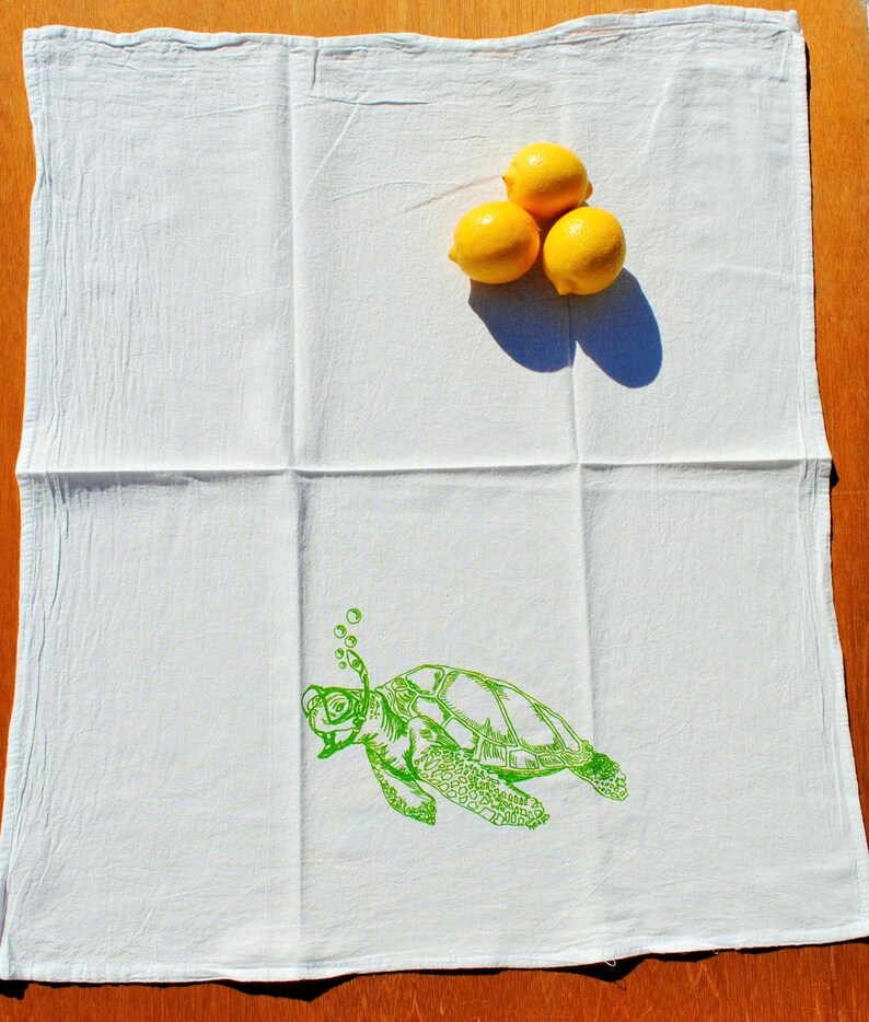 Puede incluir: Toalla de cocina blanca con una ilustraci&oacute;n verde de una tortuga marina con burbujas que salen de su nariz. Tres limones amarillos est&aacute;n en la parte superior de la toalla.